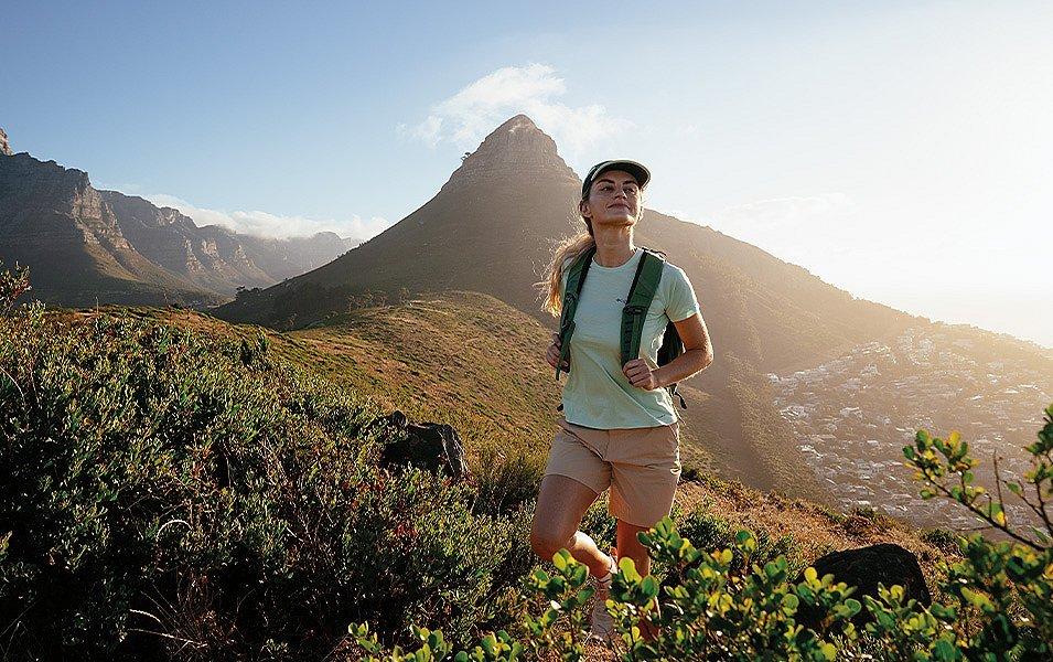 A female hiker with a backpack and sun-protective hat walks along a ridgeline after summiting a nearby peak. 