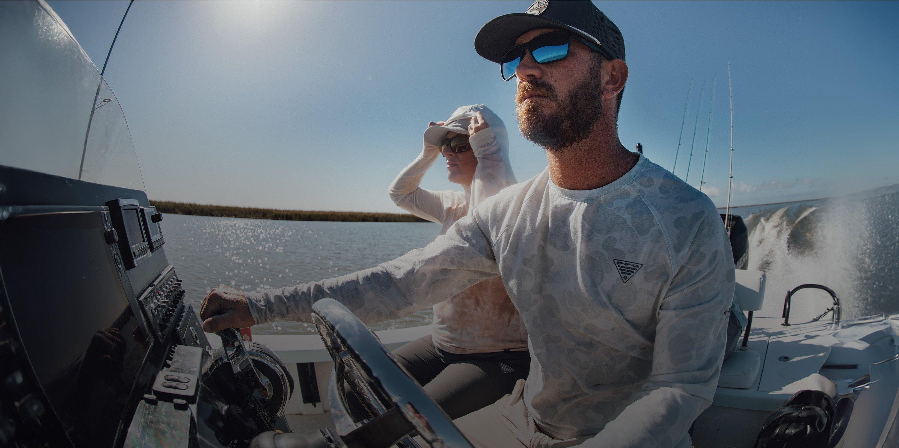 Man wearing sunglasses and cap steering a boat with another person in the background on the water.