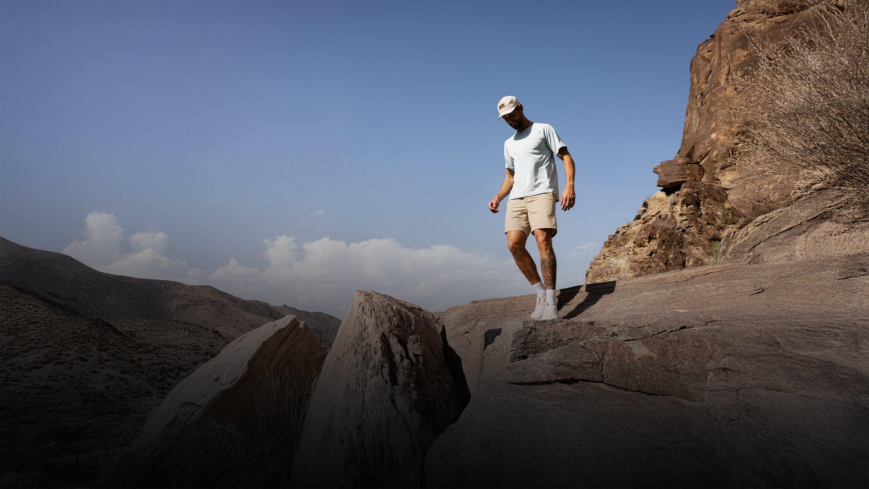 Man wearing white t-shirt, beige shorts, white cap and sneakers hiking on rocky desert terrain.