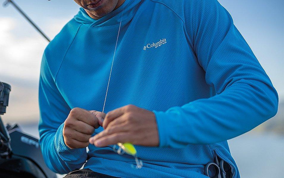 A man in a blue Columbia PFG Sun protective hoodie ties a fishing lure onto fishing line. 