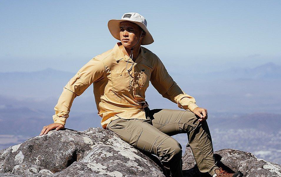 A man in Columbia Sportswear sun protective shirt, hat, and pants sits on a rock on top of a hill. 