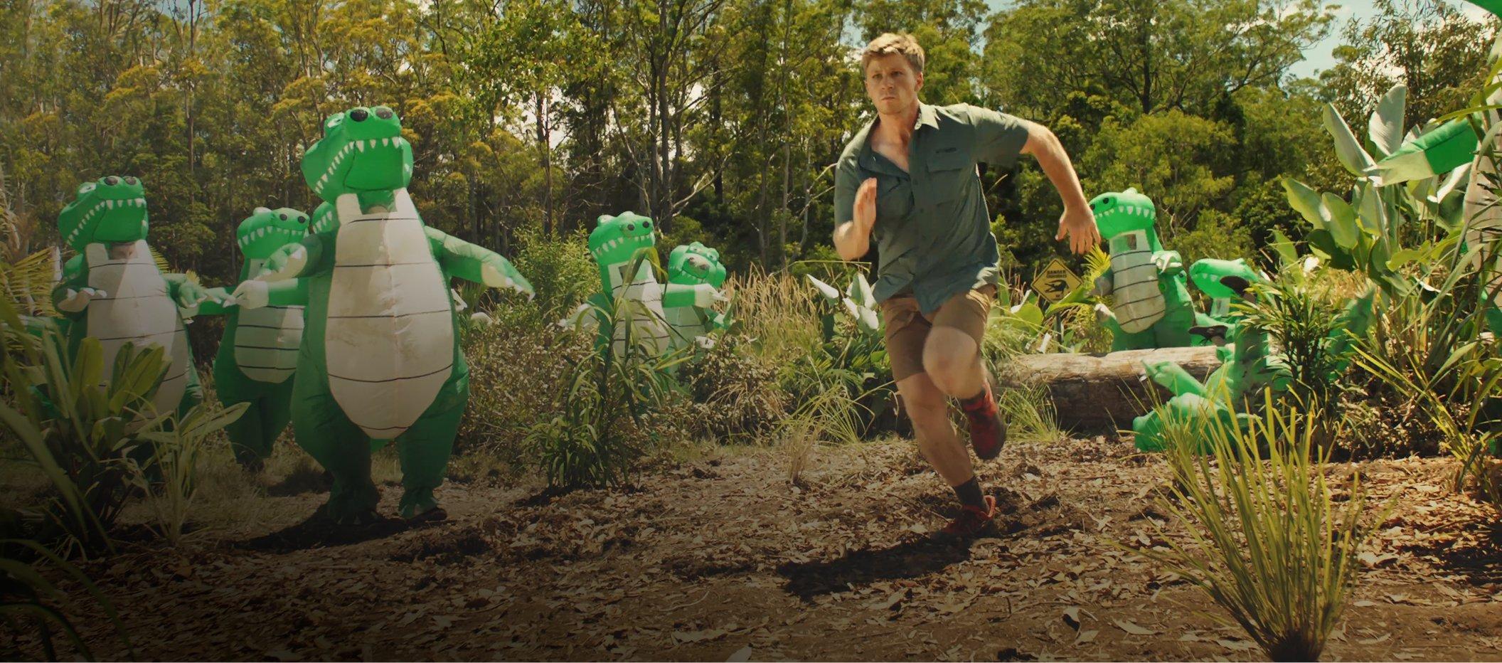 Man running through jungle setting with green toy dinosaur figures in outdoor adventure scene.