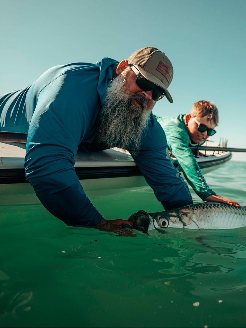 Man in blue long-sleeve shirt and cap releasing a large tarpon fish into green water from a boat.