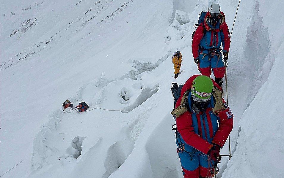 Two mountaineers, both wearing Columbia's Attack Suit, ascend a very steep section on the Khumbu Ice Field.  They are clipped into a fixed line and there are climbers crossing a crevasse field far below.   