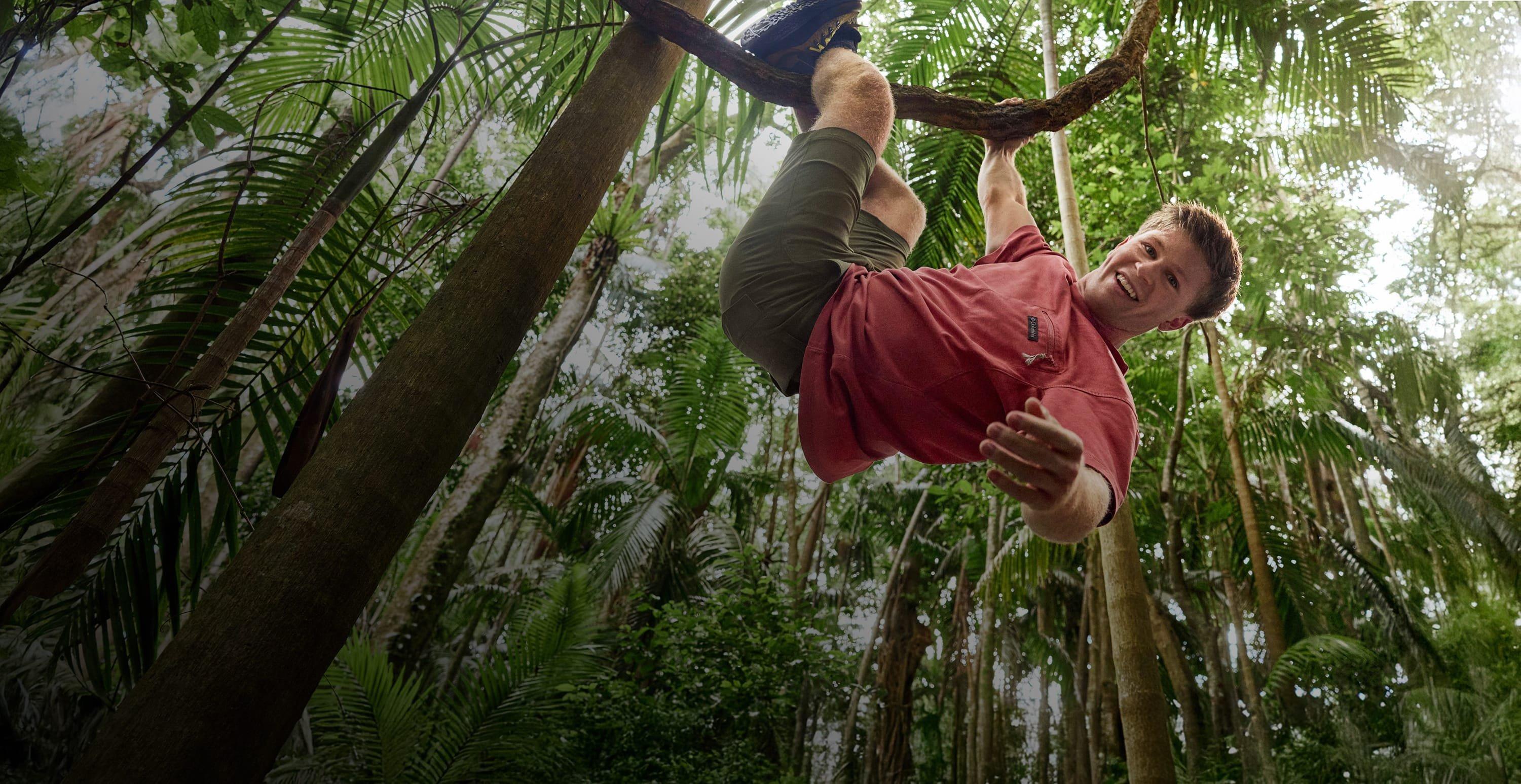 Robert Irwin climbing a tree wearing a red short sleeve shirt, green cargo shorts, and hiking boots.
