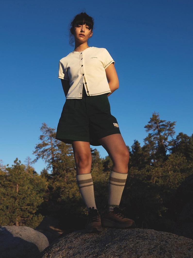 Woman standing on rock outdoors wearing cream button-up shirt, black skort, striped knee socks, and hiking boots.