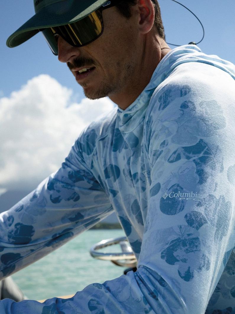 Man fishing on a boat wearing blue fish-print long sleeve shirt, green cap, and yellow-striped sunglasses.