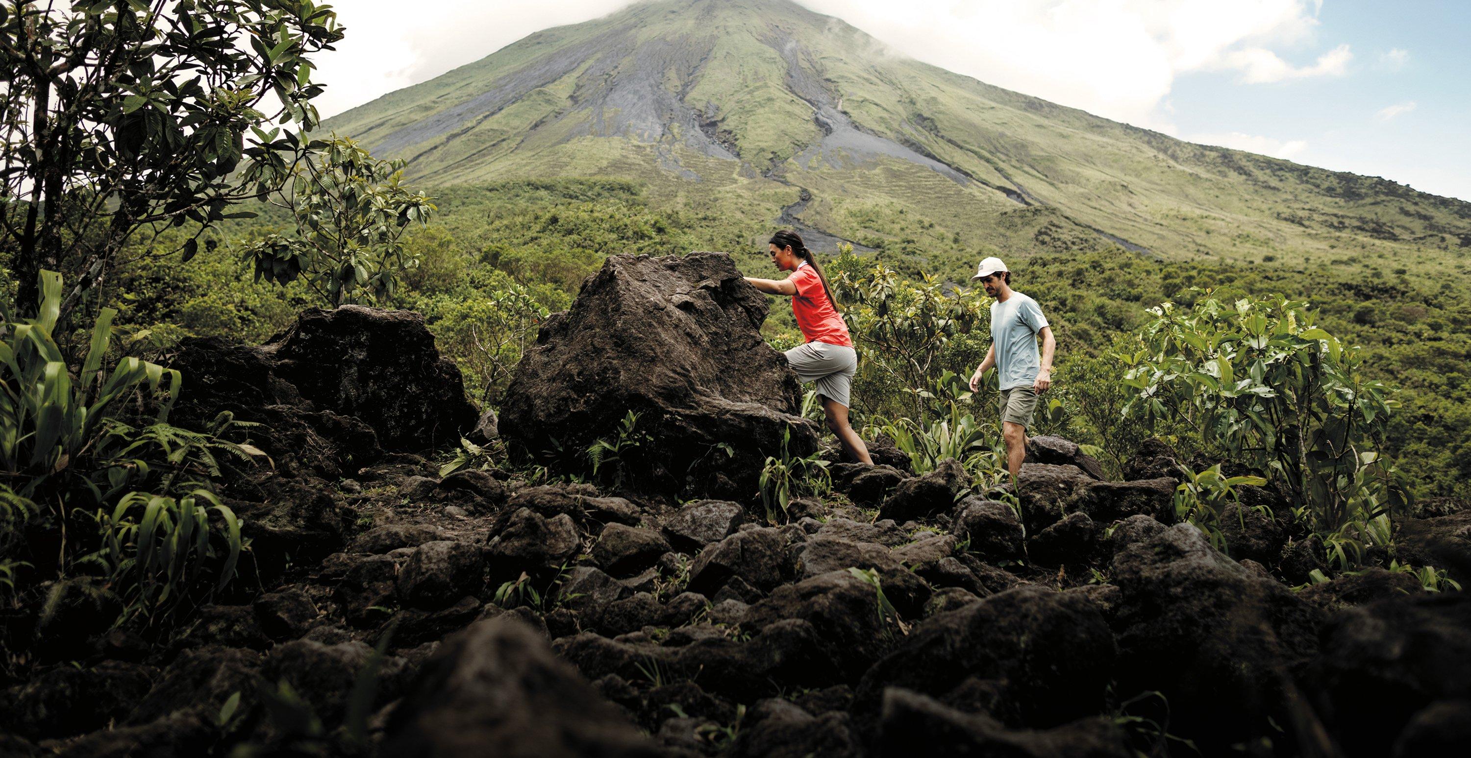 Two hikers climbing over volcanic rocks on a trail with a green mountain peak in the distance.