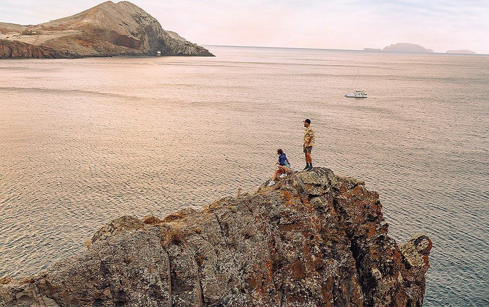 Two people enjoy a spectacular water view from their perch atop a rocky outcrop.