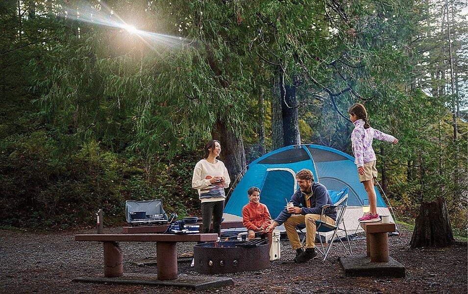 A family is enjoying each other's company as they gather around a firepit, their tent is in the background. 