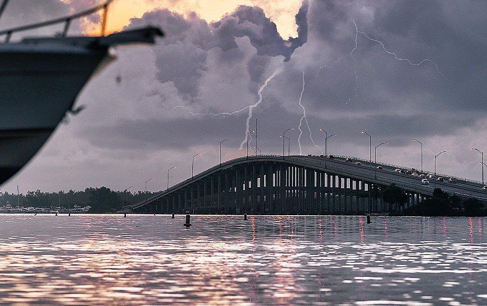 Big river with arched bridge; the evening sky is dark, with an evening lightning storm brewing in the distance. There’s a boat floating on the river the remaining rays of sun are reflecting off the water. 