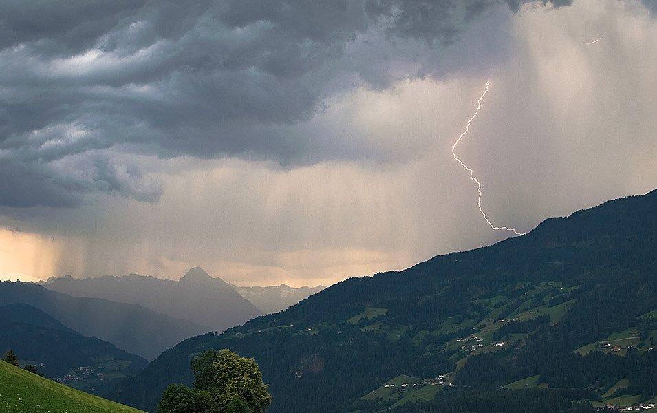 Alpine landscape with sharp vertical relief. A small village with streetlights clings to a mountain on one side of a deep valley. On the other side, there’s a green grassy field. Rain is spilling out of dark clouds and a jagged bolt of lightning is striking the hillside above the village. 

 