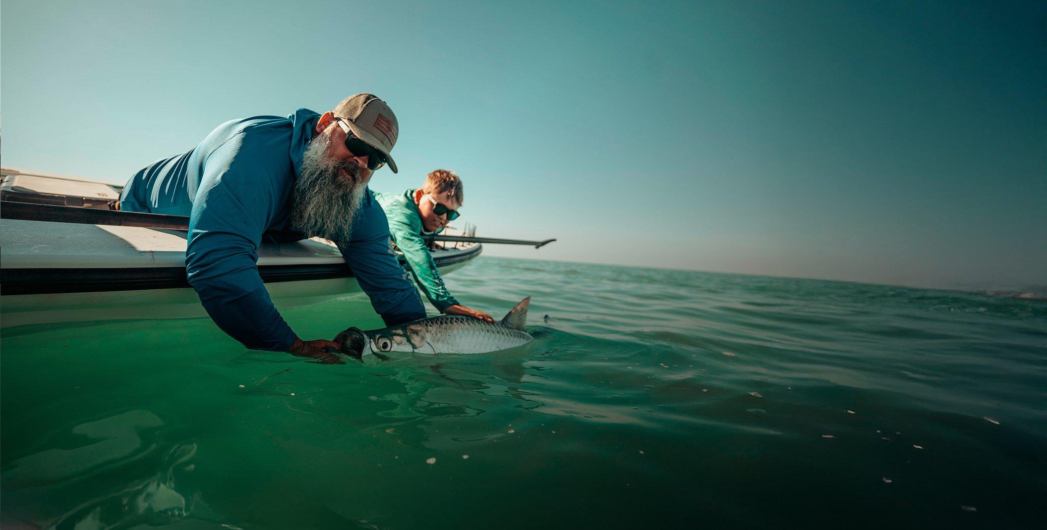 Two men releasing a large silver fish back into turquoise ocean water from the side of a boat.