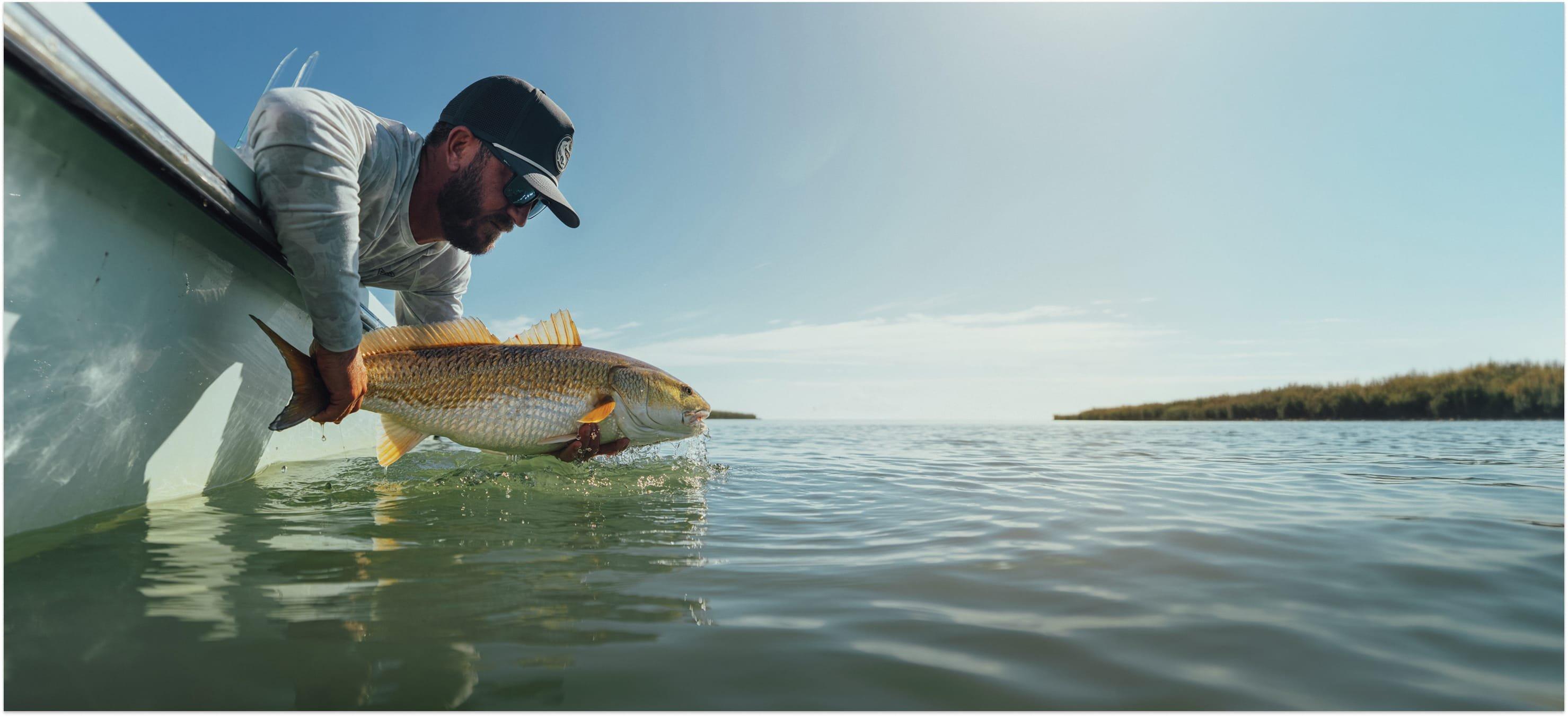 Man in cap and long-sleeve shirt releasing a redfish into calm water from the side of a boat.