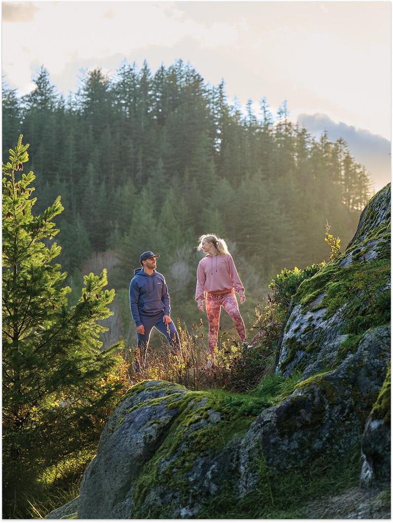 Two people hiking on a rocky, moss-covered trail surrounded by evergreen trees in natural sunlight.