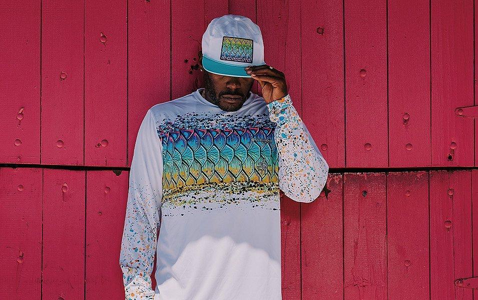 A man leans against a wooden wall, wearing a ball cap and sun-protective hoodie with distinctive Bentley graphics.