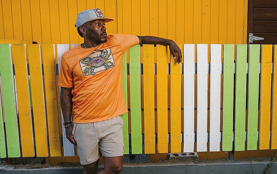 A man drapes his arm over a colorful fence, wearing a T-shirt with a Bentley crab graphic.