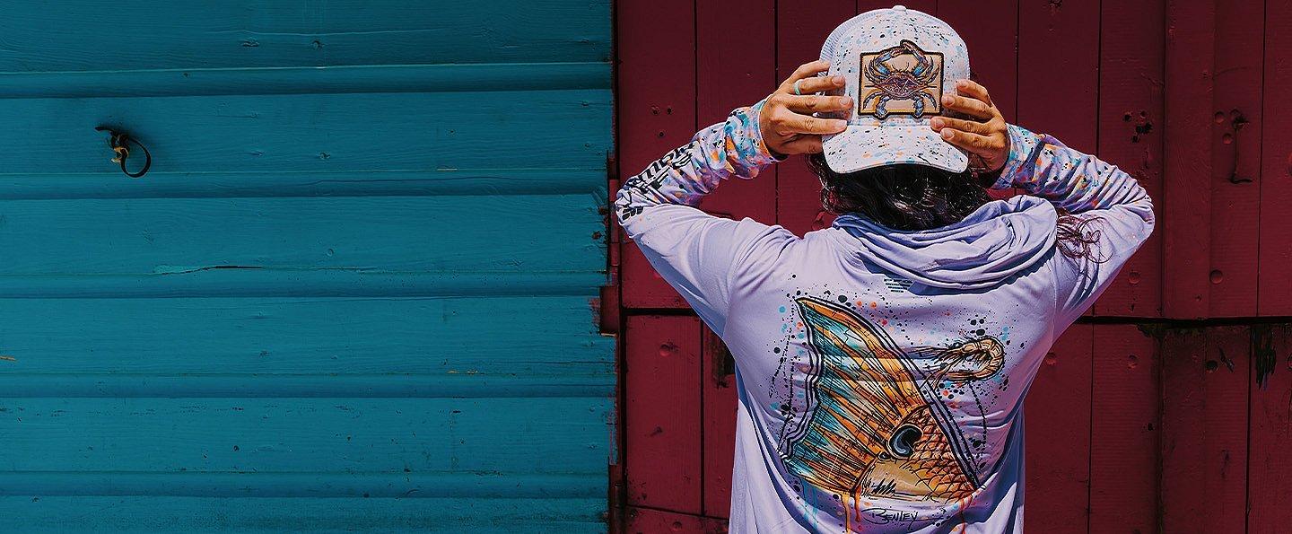 A person wears a lavender sun-protective hoodie and matching ball cap, both featuring marine life graphics. 