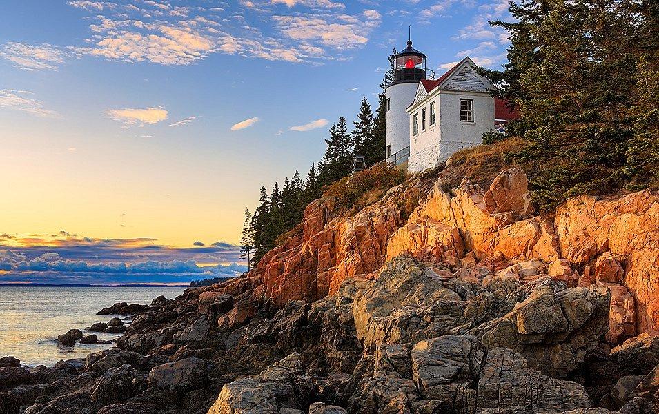 A historic lighthouse sits on a rocky bank above a big expanse of water. The lighthouse is white and you can see the warning beacon flashing.