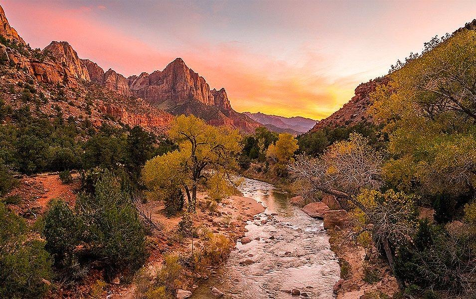  A dry, southwest landscape with banded sandstone cliffs in the background. A stream runs through the photo and the sky is turning yellow and orange.