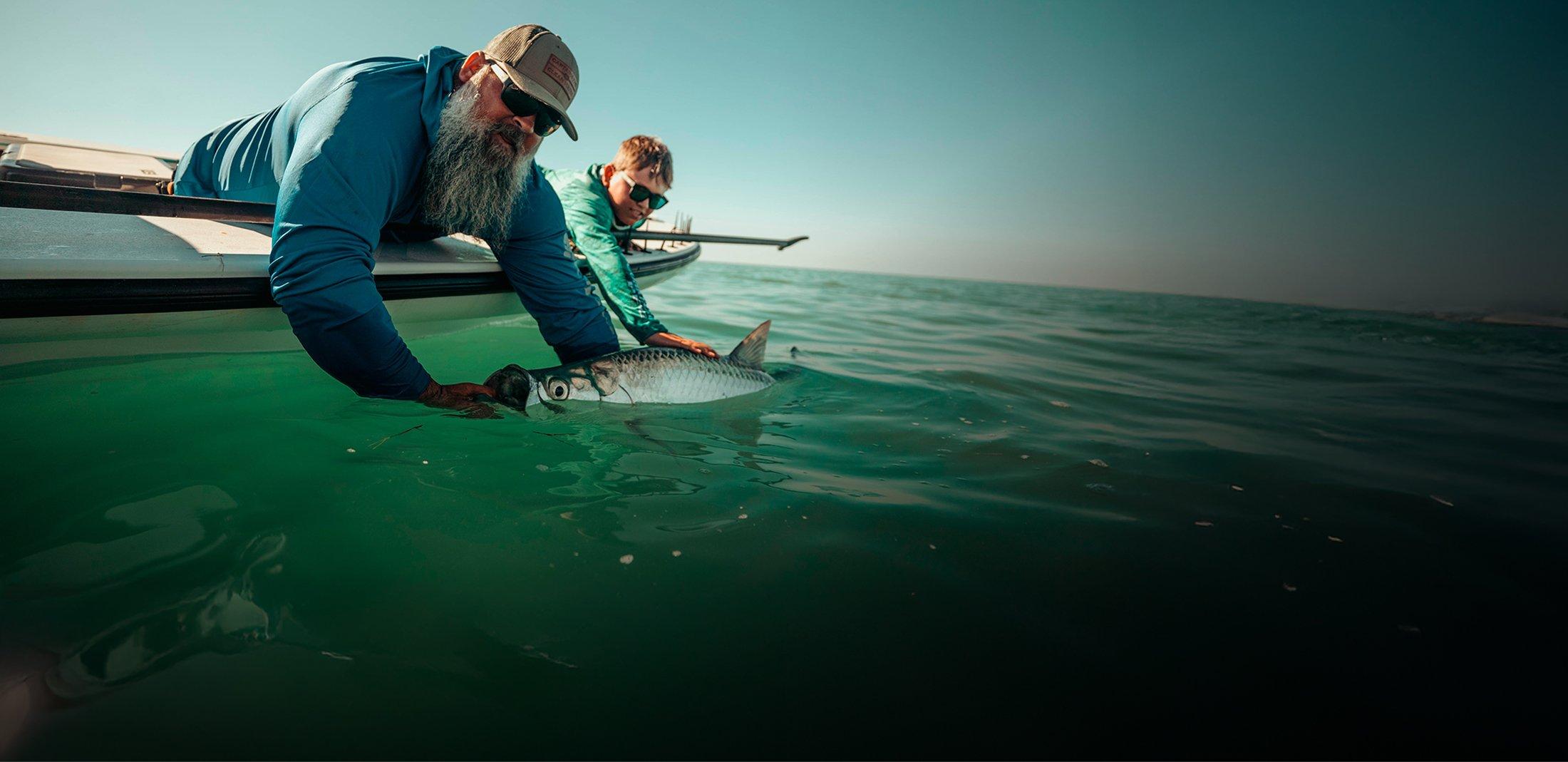 Two men in sunglasses releasing a tarpon fish into clear turquoise water from the side of a boat.