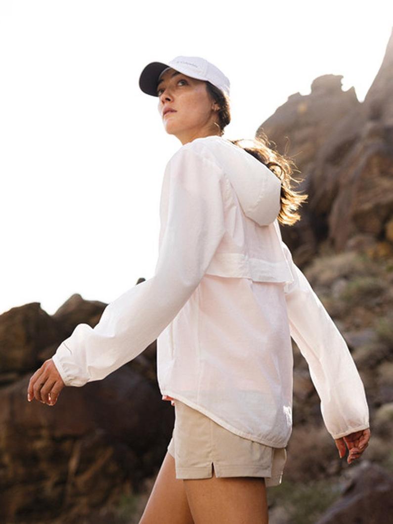 Woman wearing white cap and lightweight white jacket with beige shorts hiking outdoors on rocky terrain.