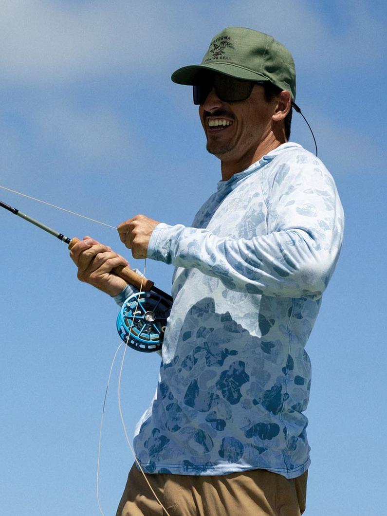 Man wearing sunglasses and green cap fly fishing in blue patterned long sleeve shirt.