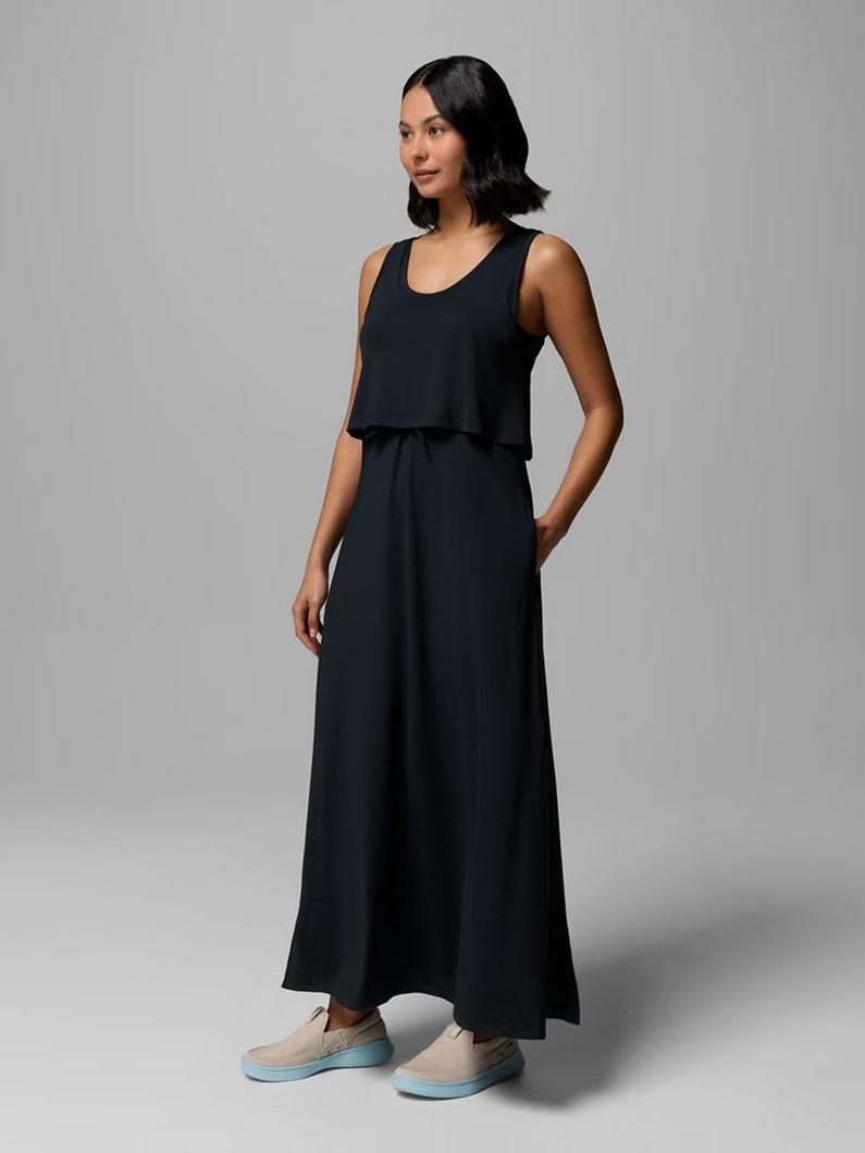 Woman wearing black sleeveless maxi dress with layered top and beige slip-on sneakers, standing.