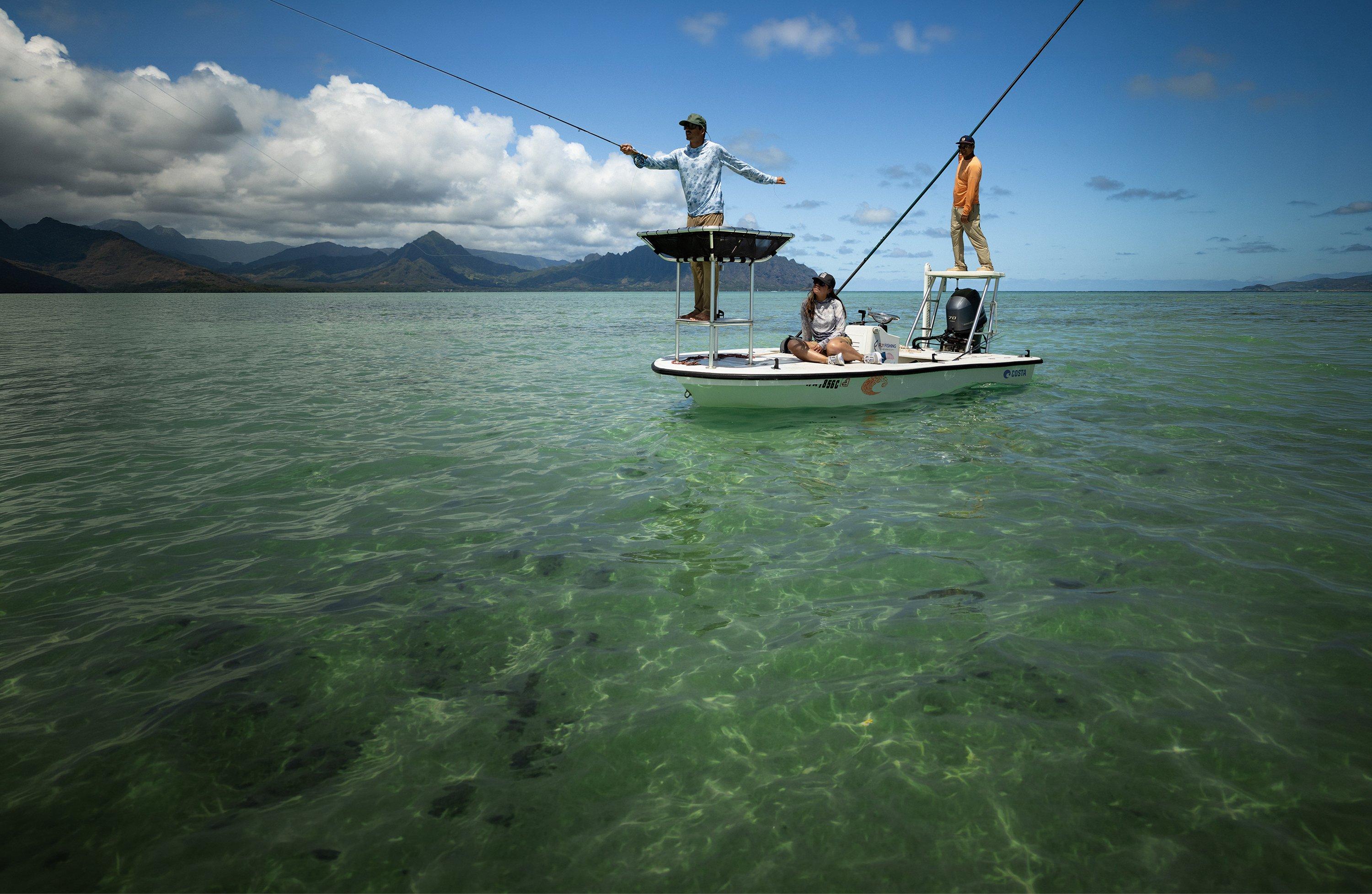 Three people fishing from a flats boat in shallow coastal waters with mountains in the distance.