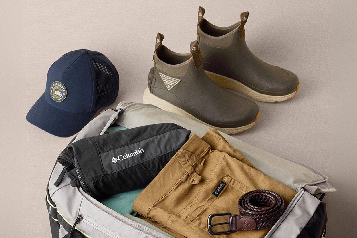 Flat lay of outdoor gear including waterproof ankle boots, navy trucker cap, duffel bag, and tan pants.