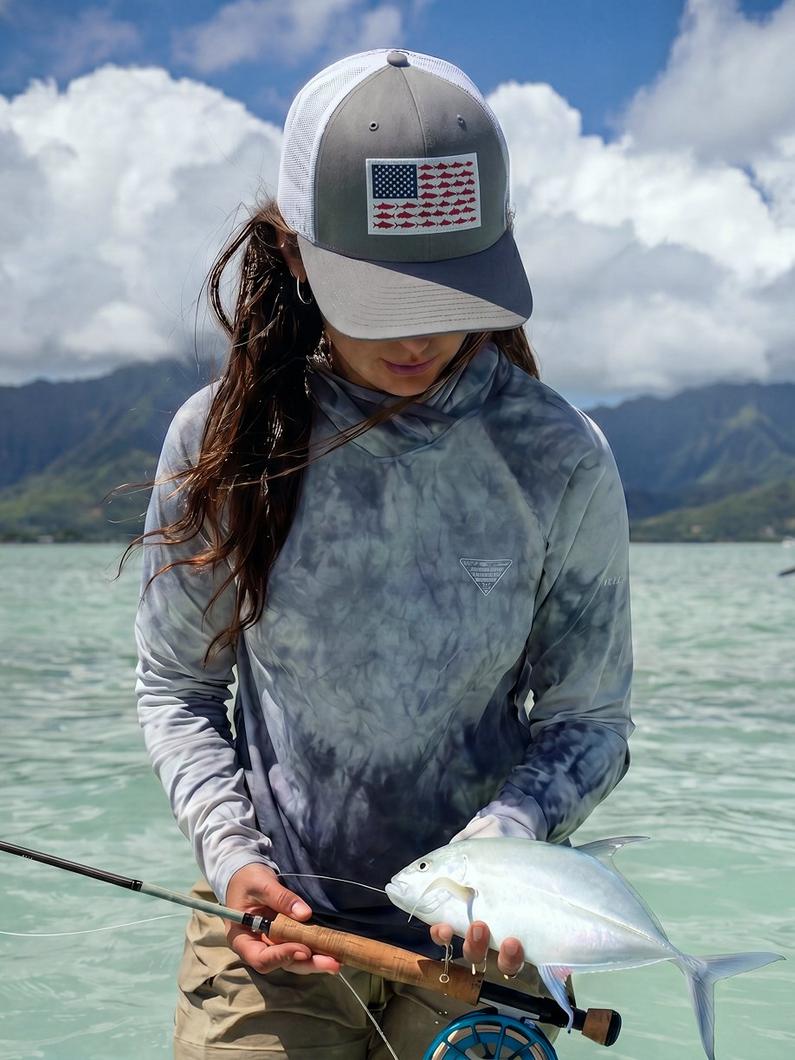 Woman wearing trucker hat with fish flag patch and tie-dye fishing shirt, holding caught fish with rod.