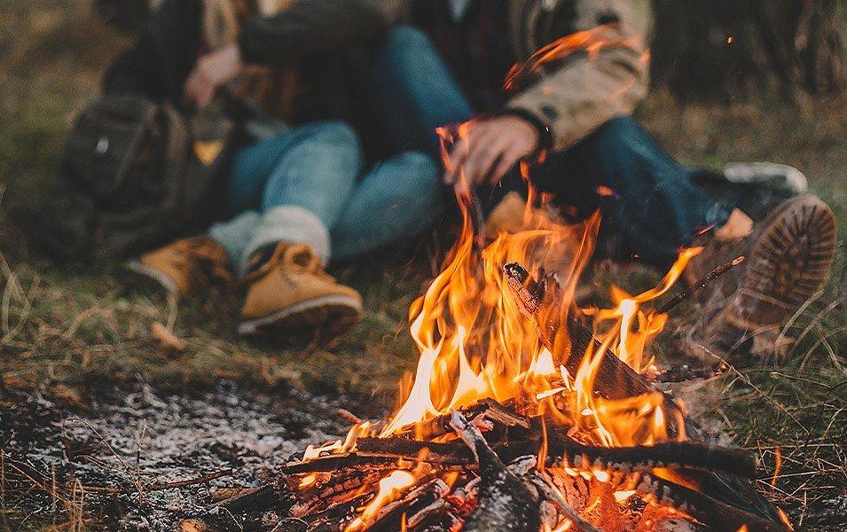 Two people, wearing Columbia Sportswear jackets and boots, sit close to each other next to a campfire. 