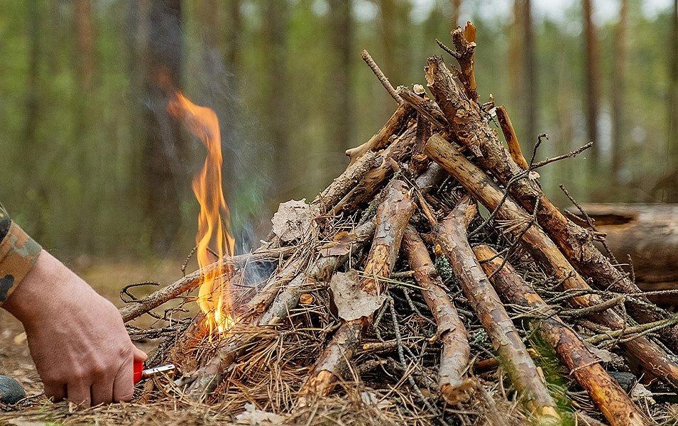 A person using a long butane lighter ignites the kindling underneath a tipi-style campfire.