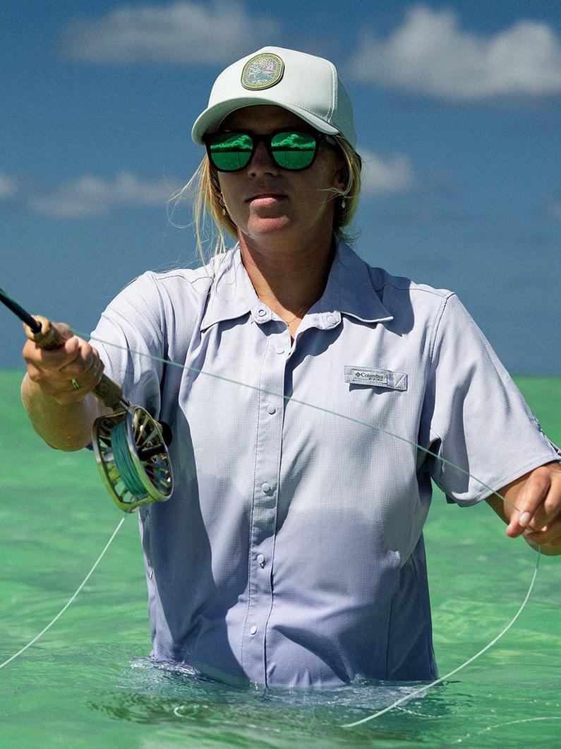 Person fly fishing in shallow water wearing white trucker cap, green-lensed sunglasses, and light blue shirt.