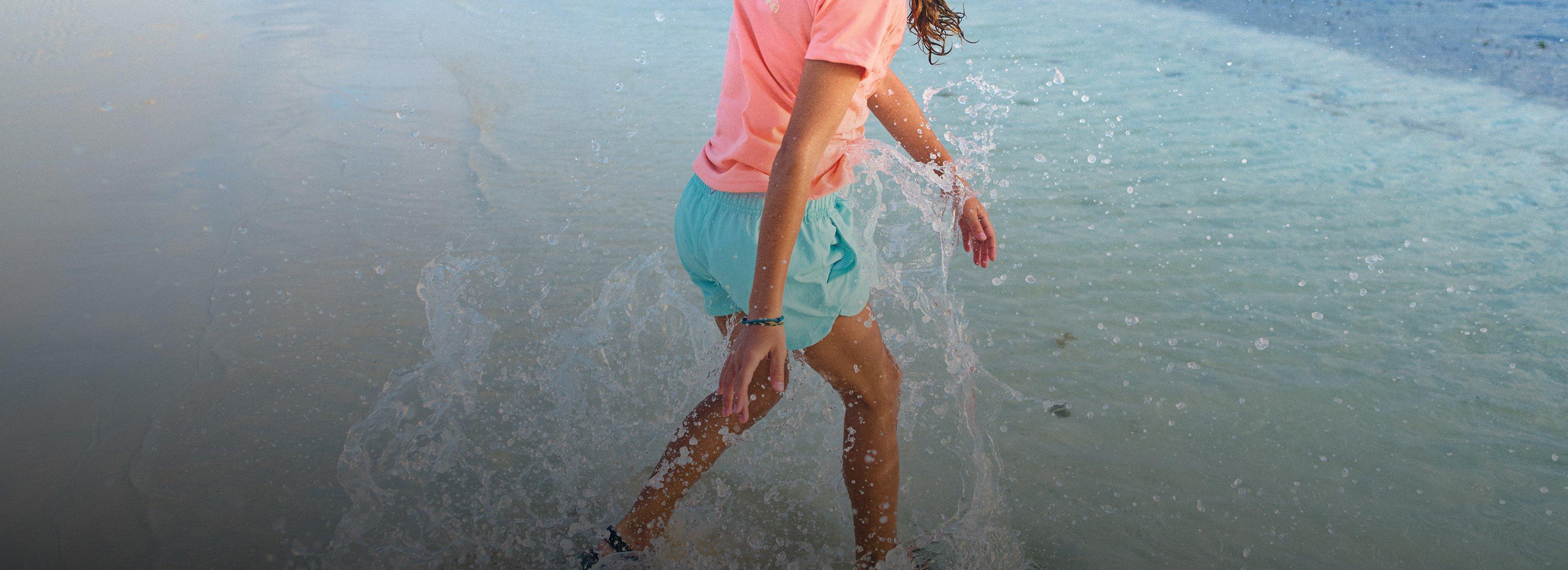 Person running through shallow ocean water wearing pink shirt and turquoise shorts, splashing playfully.