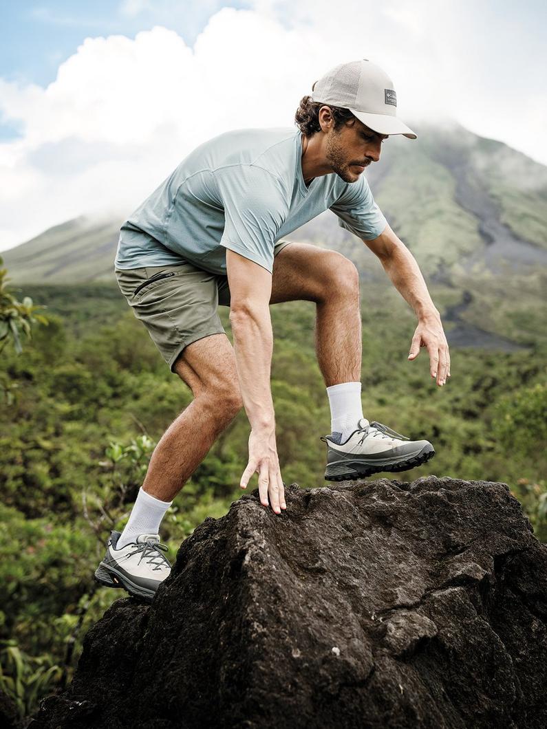 Man hiking over rocks wearing light blue shirt, olive shorts, white cap, and gray hiking shoes.