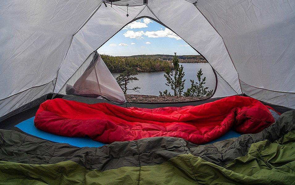 Inside a tent you can see two sleeping bags positioned on sleeping pads. The tent fly is open, with a view of a lake and trees outside.