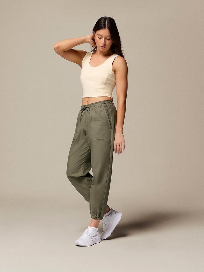 Woman wearing cream crop top and olive green jogger pants with white sneakers, hand touching hair.