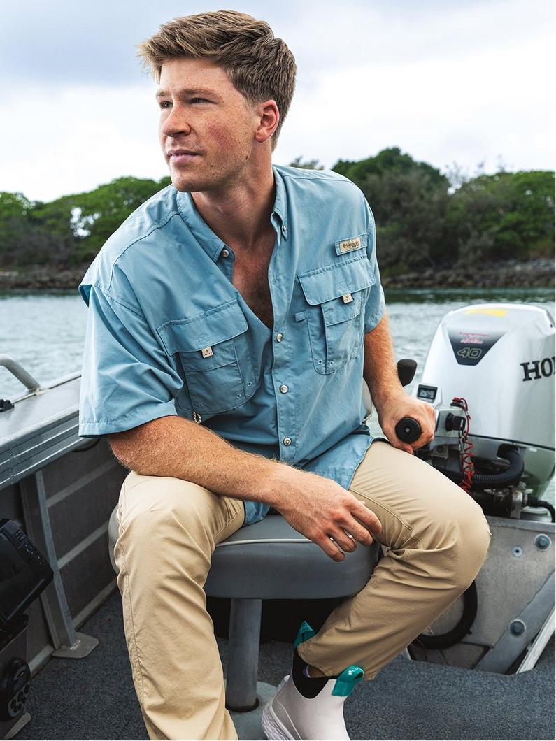 Man sitting on boat wearing light blue short-sleeved fishing shirt with chest pockets and khaki pants.