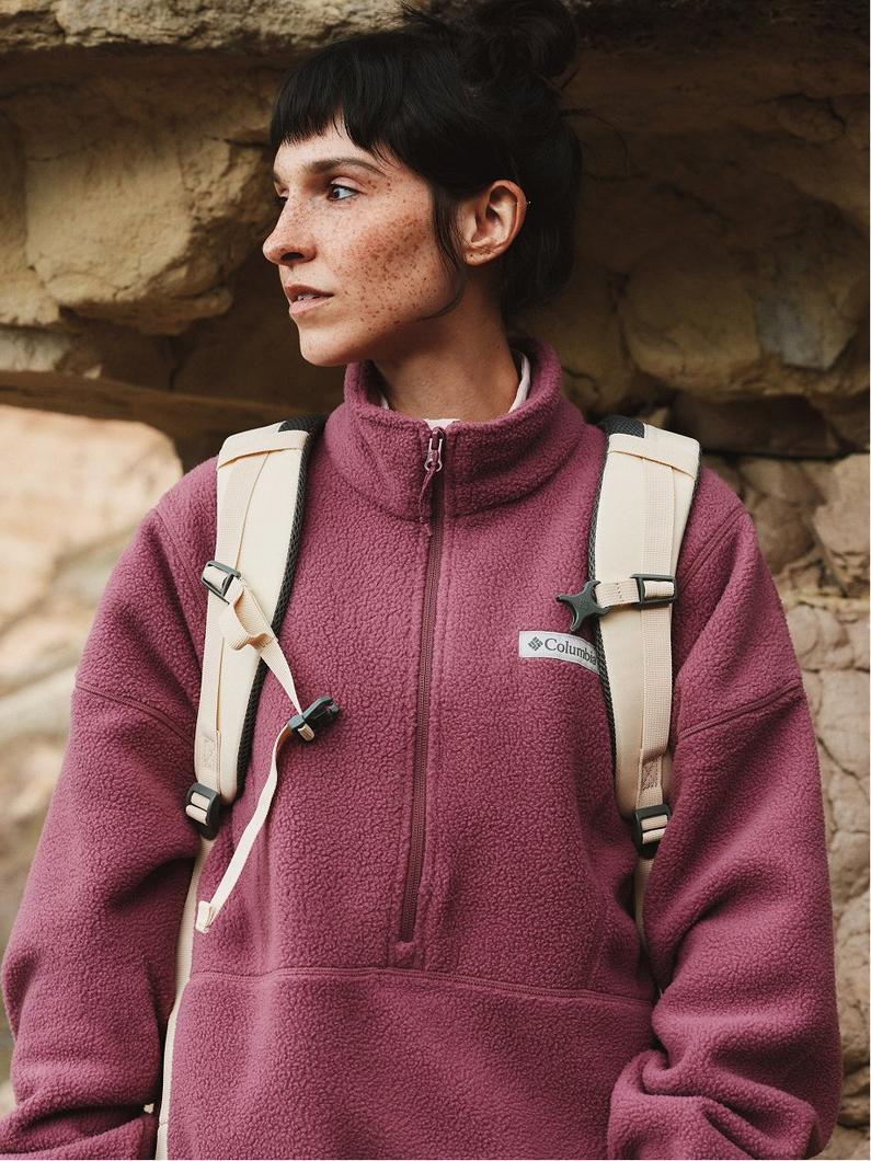 Person wearing burgundy fleece pullover with half-zip closure and beige backpack, outdoors near rock wall.
