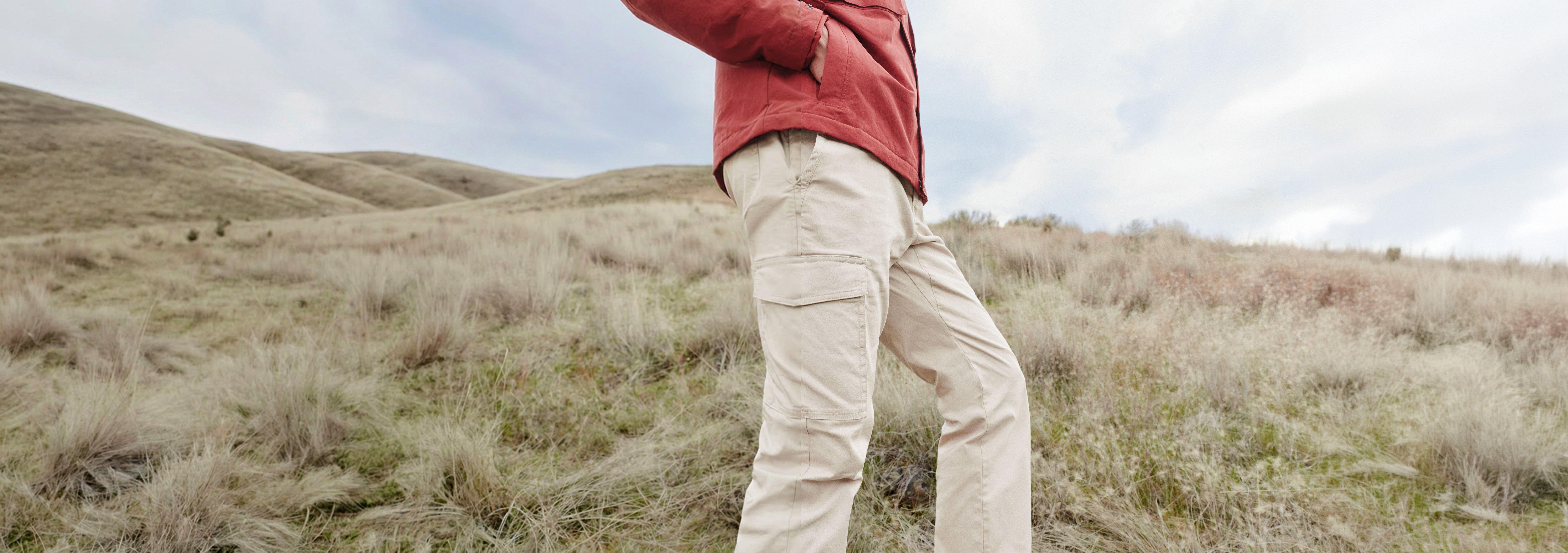 Person wearing beige cargo pants and coral jacket hiking across grassy hillside terrain.