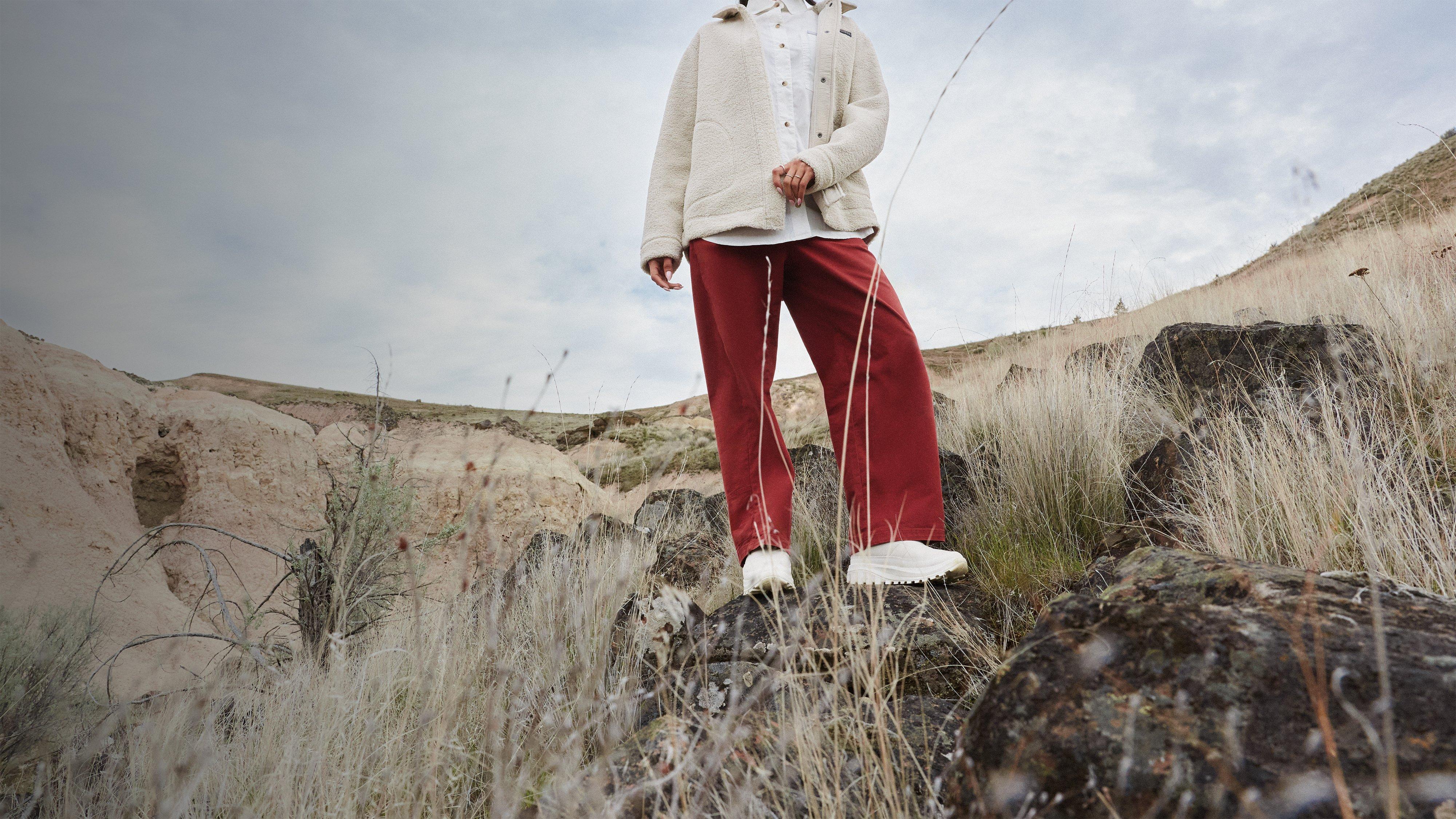 Person standing on rocky terrain wearing cream fleece jacket, burgundy joggers, white sneakers, and black cap.