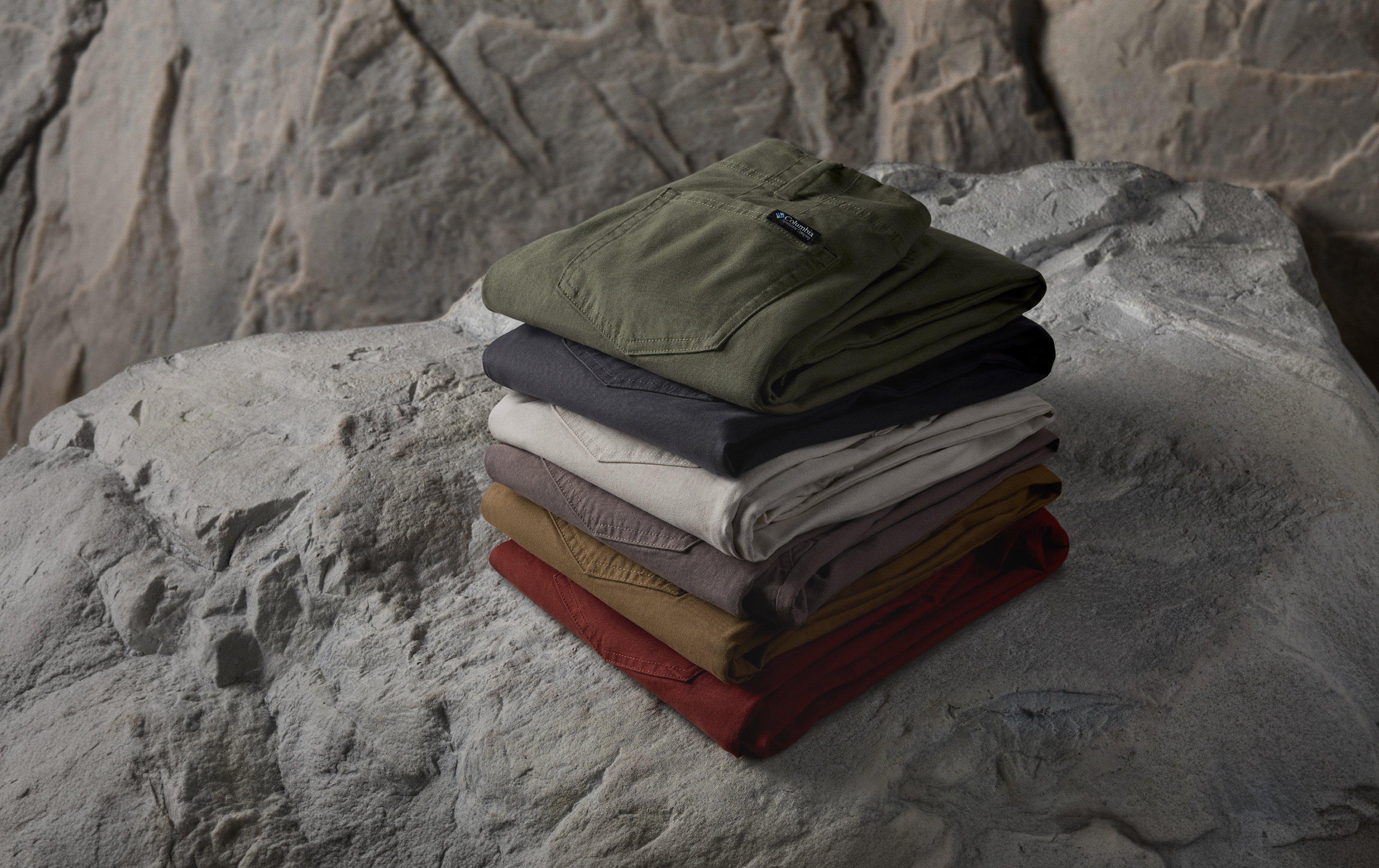 Stack of folded pants in various colors including olive green, black, cream, tan, and red plaid on stone surface.