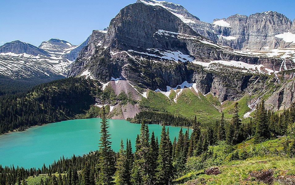 A pristine forest surrounds a round glacial lake at the base of a snow-covered granite peak.