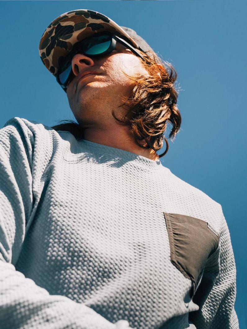 Person wearing white textured sweater with chest pocket, camouflage cap, and sunglasses outdoors.