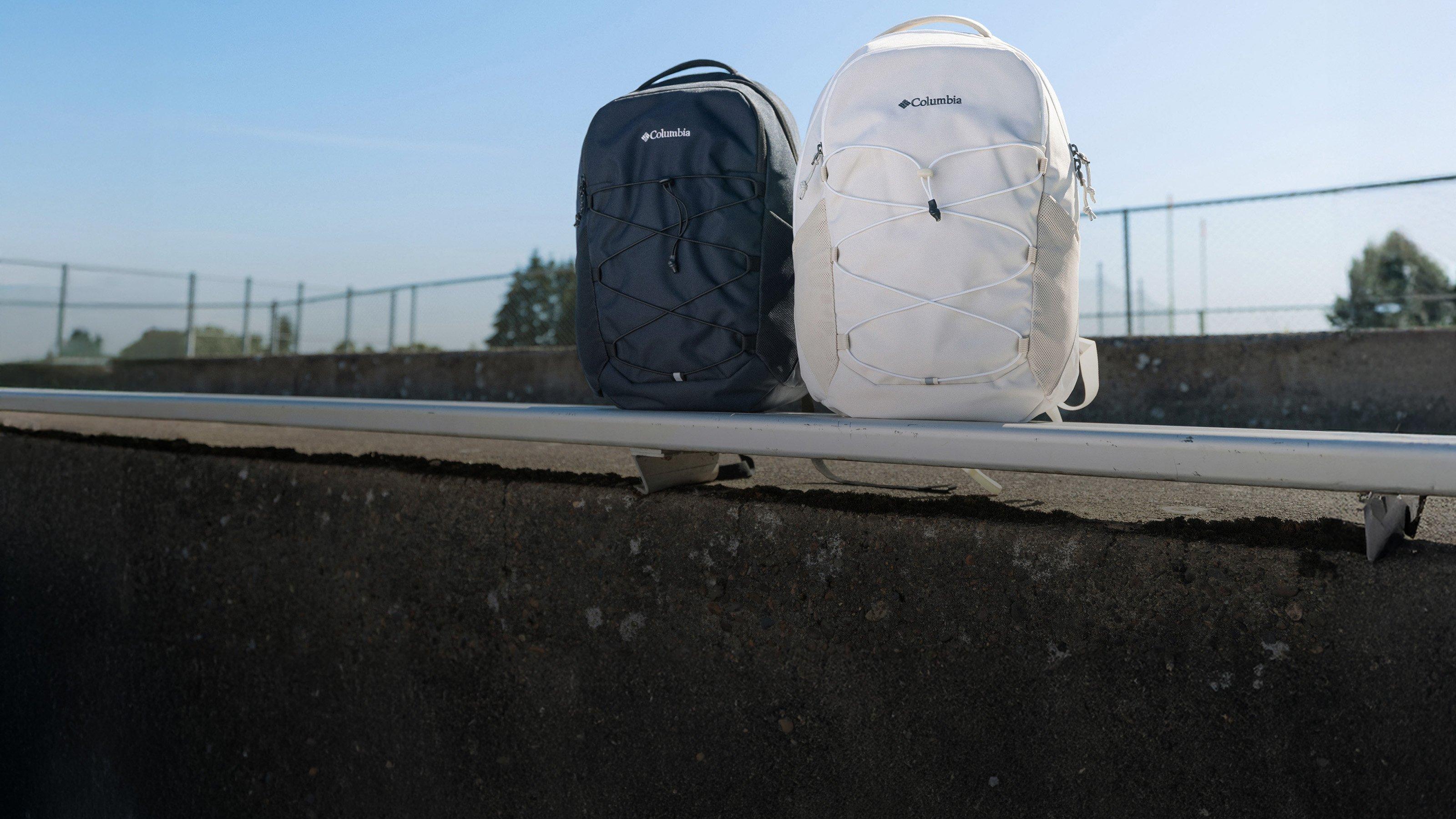 Two backpacks with elastic cord detailing on front, one in navy and one in cream, sitting on a bench.