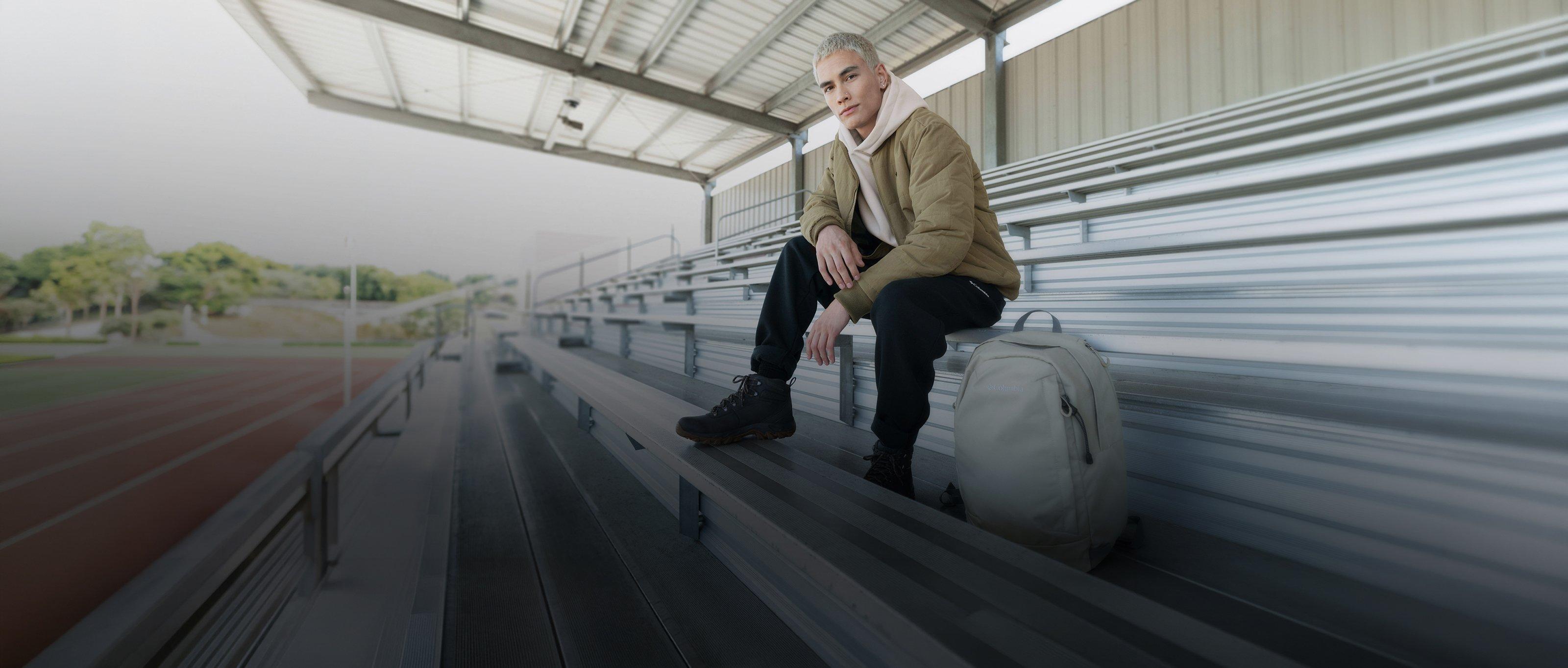 Person sitting on stadium bleachers wearing olive jacket and black pants with gray backpack beside them.