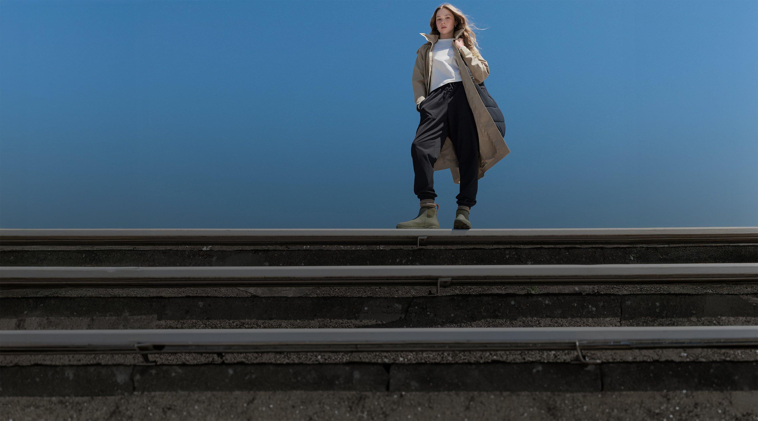 Woman standing on concrete steps wearing tan trench coat, white top, black pants, and beige boots.