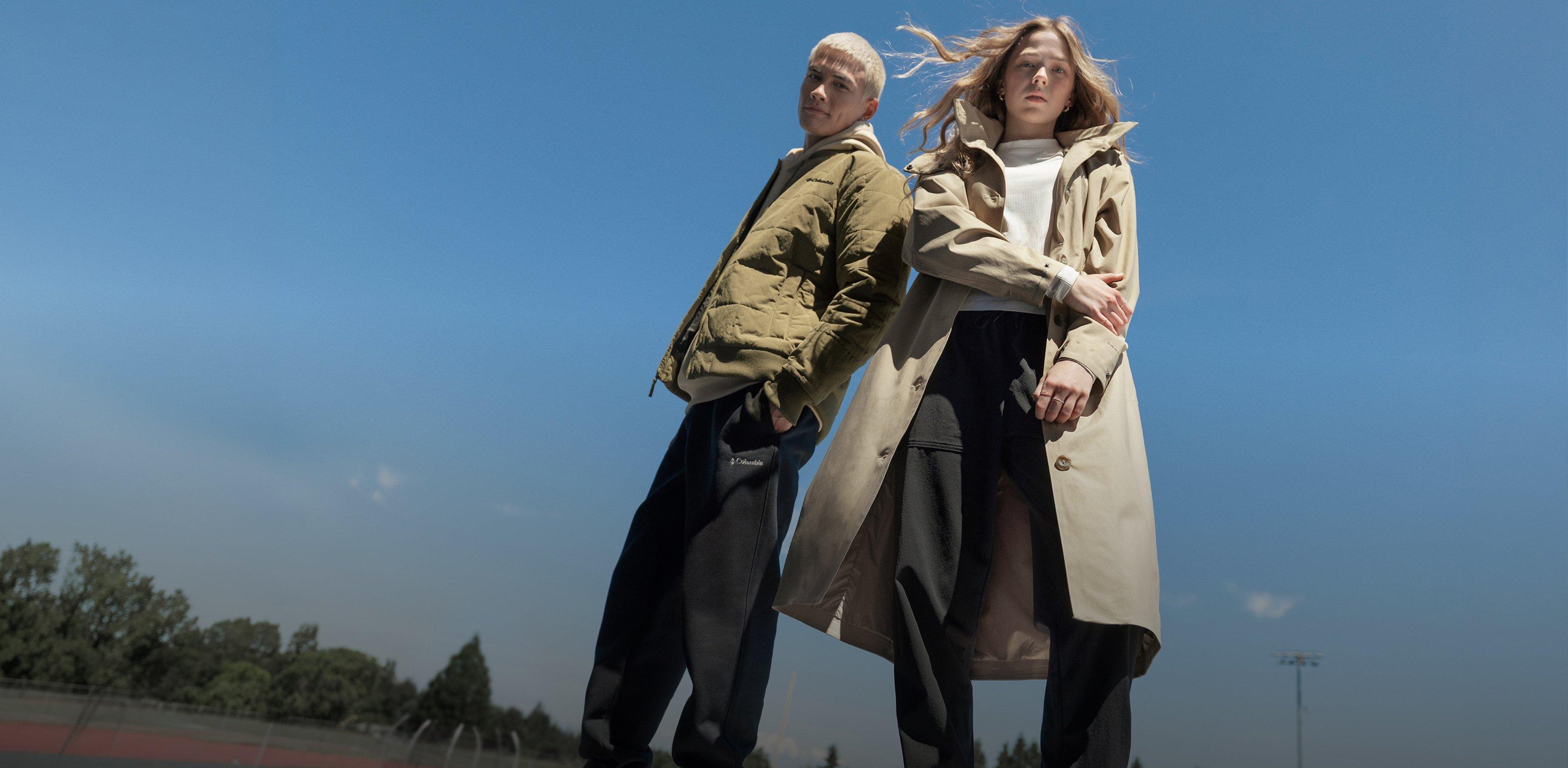 Two models standing back-to-back wearing hooded jackets and dark pants outdoors on a clear day.