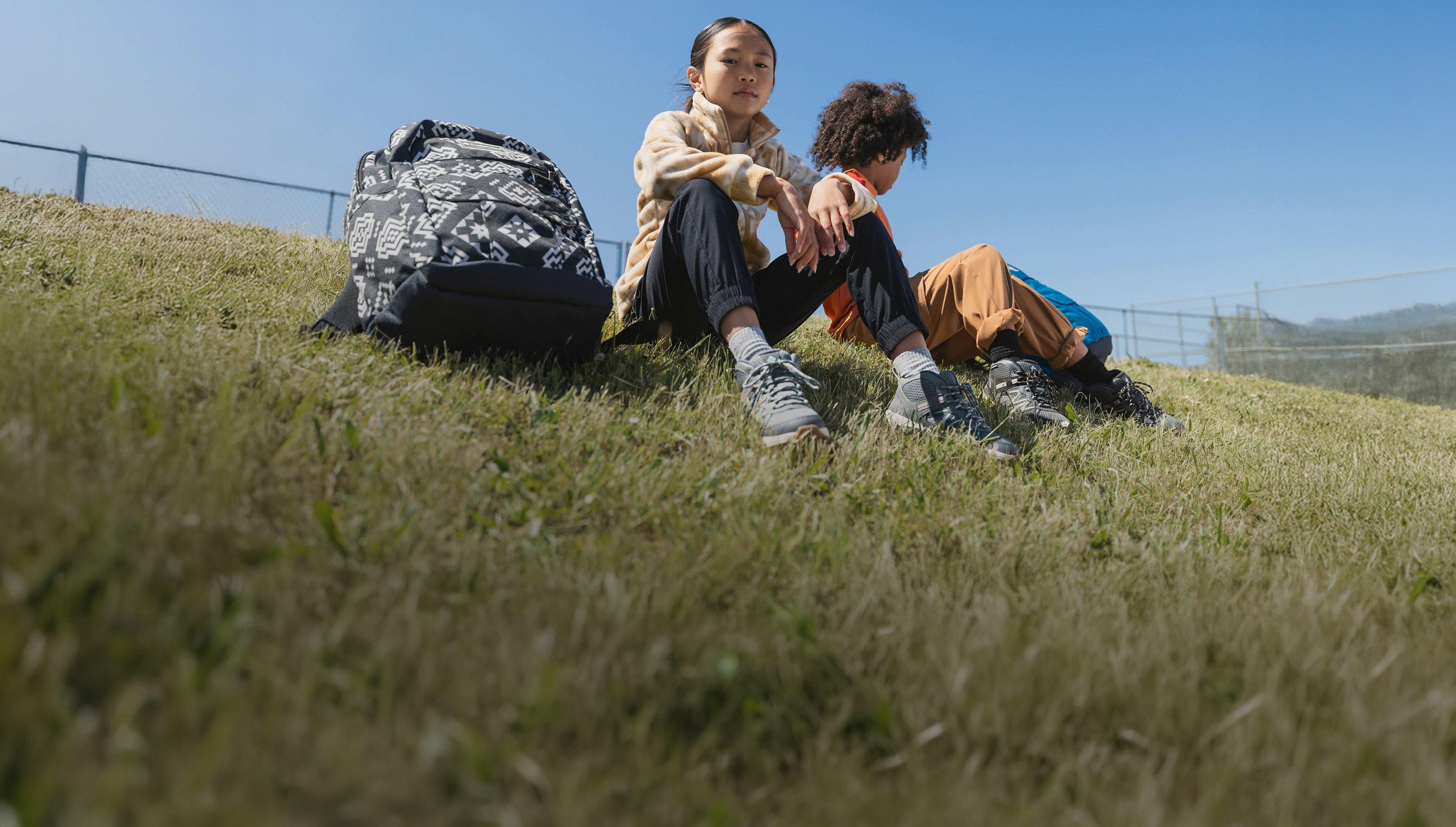 Two children sitting on grassy hillside wearing sneakers and casual clothing with a patterned backpack nearby.